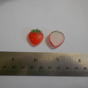 May include: Two strawberry-shaped resin charms. One is a whole strawberry with red and green details, the other is a cross-section showing the white interior. A ruler is placed next to the charms for scale.