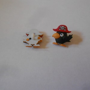 Pode incluir: Dois botões de plástico, um é um fantasma branco com detalhes laranja e o outro é um pássaro preto que usa um chapéu de pirata vermelho com uma caveira e ossos cruzados.