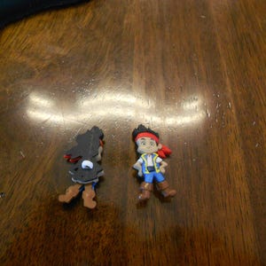 Può includere: Due piccole e colorate statuette di pirati dei cartoni animati. Una è una silhouette nera con accenti rossi, e l'altra è un pirata ragazzo con una fascia rossa, un gilet blu e stivali marroni. Sono su una superficie di legno scuro.