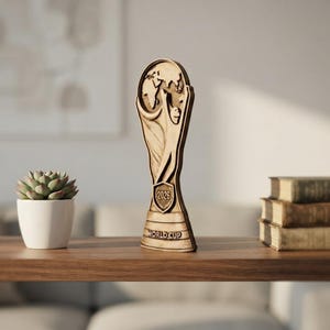 May include: A tan-colored, flat-profile trophy featuring a stylized figure holding up a globe. The trophy includes a shield-shaped emblem with the text 2026 and a base with the text WORLD CUP embossed in a dark, weathered finish.