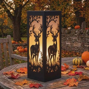 Peut inclure: Une lanterne rectangulaire noire avec un motif découpé représentant une silhouette de cerf sur chaque face. La lanterne émet une lumière chaude et éclatante de l'intérieur, illuminant les motifs forestiers complexes sur le cadre sombre.
