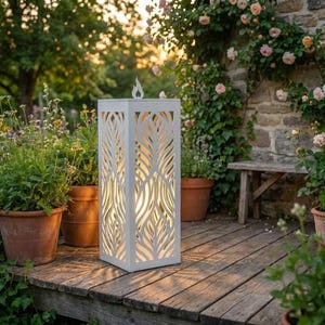 Può includere: Una lanterna alta e quadrata in metallo bianco con intricati ritagli simmetrici a forma di foglia sui lati. La lanterna emette una luce calda dall'interno ed è sormontata da un piccolo elemento decorativo a forma di fiamma al centro.