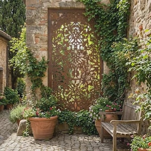Puede incluir: Un panel metálico rectangular alto con acabado color óxido y un intrincado patrón floral simétrico cortado con láser. El panel está colocado contra una pared de piedra, flanqueado por hiedra trepadora y plantas en macetas sobre un camino empedrado.