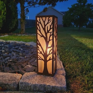 Può includere: Una lanterna rettangolare da esterno con una cornice marrone scuro caratterizzata da un design a silhouette di albero tagliato al laser. La lanterna emette un bagliore caldo e soffuso dall'interno, illuminando l'intricato motivo di rami e tronco.