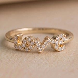 Könnte beinhalten: Ein goldener Ring mit dem Wort LOVE in Großbuchstaben auf der Vorderseite. Die Buchstaben sind mit kleinen, klaren, funkelnden Steinen besetzt, die die gesamte Oberfläche jedes Buchstabens bedecken und ein helles und reflektierendes Aussehen erzeugen.