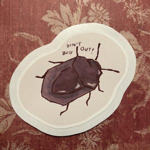 Könnte beinhalten: Ein gestanzter Aufkleber mit einer dunkelvioletten, schildförmigen Käferillustration auf hellcremefarbenem Hintergrund. Der Text "DON'T BUG OUT!" ist in schwarzer Tinte über dem Insekt geschrieben, das sechs dünne Beine und zwei Fühler hat.