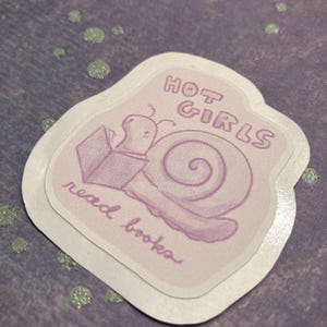 Puede incluir: Una pegatina troquelada de color morado claro que muestra un caracol de dibujos animados leyendo un libro abierto. El texto "HOT GIRLS" está impreso en una fuente negrita y redondeada sobre el caracol, y "read books" en cursiva debajo.