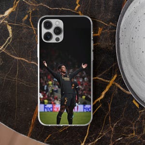 Puede incluir: Una funda transparente para tel&eacute;fono con un estampado gr&aacute;fico de un jugador de f&uacute;tbol con camiseta negra, el n&uacute;mero 5 y el texto Emirates Fly Better, de pie en un campo verde con los brazos extendidos frente a una multitud de estadio borrosa.