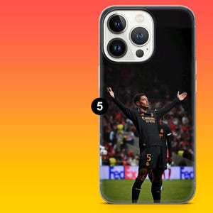Puede incluir: Una funda transparente para smartphone con la imagen impresa de un futbolista vistiendo una camiseta negra con el n&uacute;mero 5 en el pantal&oacute;n corto, de pie en un campo con los brazos extendidos. El jugador est&aacute; frente a un fondo de estadio.