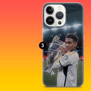 Puede incluir: Una funda transparente para smartphone con la imagen impresa de un futbolista con camiseta blanca sosteniendo un gran trofeo plateado en un campo. El tel&eacute;fono tiene un sistema de c&aacute;mara de triple lente y un icono negro con el n&uacute;mero 3.