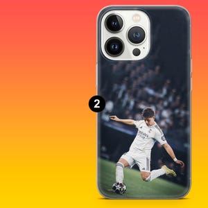 May include: A clear smartphone case featuring a printed graphic of a soccer player in a white uniform kicking a soccer ball on a field. The case has a cutout for a triple-lens camera system on the back, with a black number 2 icon on the left side.