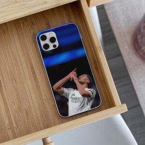 Puede incluir: Una funda rectangular para smartphone con una impresi&oacute;n fotogr&aacute;fica de un jugador de f&uacute;tbol con camiseta blanca y el texto Emirates Fly Better, haciendo un gesto con la mano mientras mira hacia arriba sobre un fondo de estadio azul borroso.