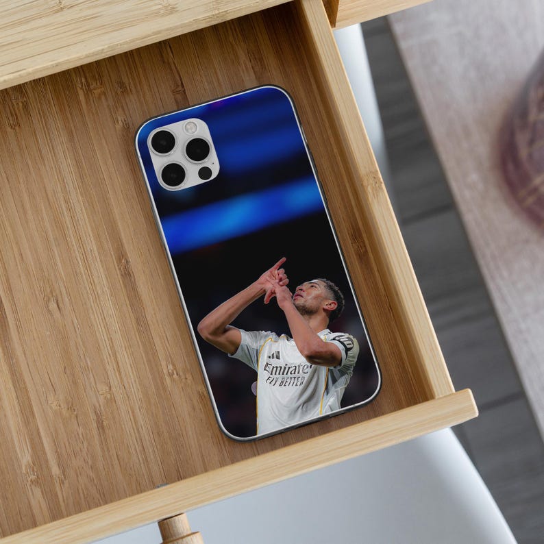Puede incluir: Una funda rectangular para smartphone con una impresi&oacute;n fotogr&aacute;fica de un jugador de f&uacute;tbol con camiseta blanca y el texto Emirates Fly Better, haciendo un gesto con la mano mientras mira hacia arriba sobre un fondo de estadio azul borroso.