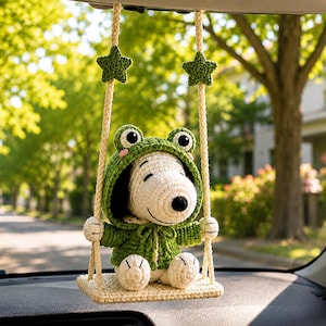 Può includere: Una statuetta di Snoopy all'uncinetto che indossa una felpa verde a tema rana con guance rosa, seduta su una piccola altalena rettangolare beige sospesa da due corde color crema decorate con due piccole stelle verdi all'uncinetto.