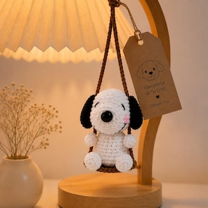 May include: A white and black crocheted dog figure sitting on a small brown swing. The figure has a black nose, black ears, and pink cheeks. A brown paper tag hangs from the swing, featuring a dog illustration and the text HANDMADE WITH LOVE THANK YOU.