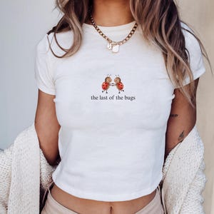 May include: A white short-sleeved t-shirt featuring a graphic of two ladybirds holding hands with the text "the last of the bugs" printed underneath. The shirt has a crew neckline and is paired with a gold chain necklace with a small pendant.