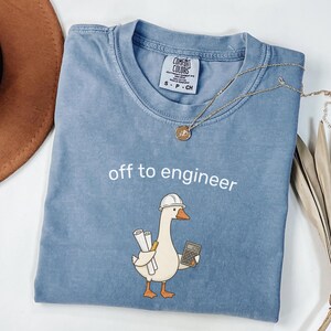 Może przedstawiać: Niebieski t-shirt z krótkim rękawem z grafiką białej gęsi w kasku ochronnym, trzymającej plany i kalkulator. Nad gęsią znajduje się napis "off to engineer". Koszulka posiada metkę marki Comfort Colors przy kołnierzu.