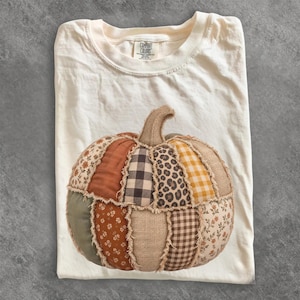 May include: A cream-colored t-shirt featuring a large, central pumpkin graphic made of various fabric patches. The pumpkin design includes sections of burlap, leopard print, black and white buffalo check, orange gingham, and floral patterns with frayed edges.