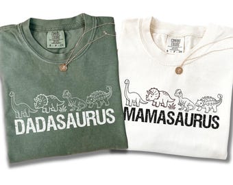 Könnte beinhalten: Ein salbeigrünes T-Shirt mit weißen Dinosaurier-Grafiken und dem Text DADASAURUS sowie ein weißes T-Shirt mit schwarzen Dinosaurier-Grafiken und dem Text MAMASAURUS. Beide T-Shirts haben einen Rundhalsausschnitt und ein Comfort Colors Etikett.