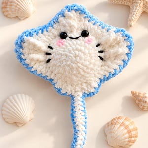 May include: A crocheted stingray plush toy featuring a white body with a light blue border, black plastic eyes, a stitched black mouth, and pink embroidered cheeks. The stingray has a long, textured tail and is surrounded by seashells.