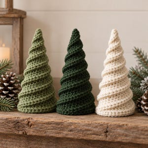 May include: Three cone-shaped decorative items crafted from yarn in a spiral crochet pattern. The set includes one light olive green tree, one dark forest green tree, and one cream-coloured tree, each featuring a textured, ribbed surface.
