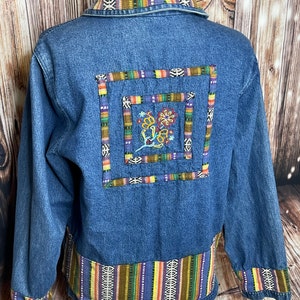 May include: A blue denim jacket featuring a collar, cuffs, and a bottom hem trimmed with a multi-colored striped fabric. The back displays a square patch with a central embroidered floral design, framed by two concentric borders of the same striped fabric.