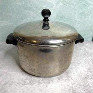 May include: A stainless steel pressure cooker with a lid and black handle. The lid has a black knob. The pot is cylindrical and has two black handles on the sides. The pot is sitting on a gray surface.