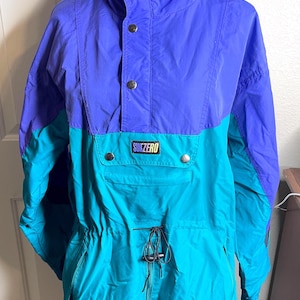May include: A color-blocked pullover jacket featuring a purple upper section and a teal lower section. The jacket has a high collar, a front placket with silver-tone snap buttons, a central pocket with a SUBZERO patch, and a drawstring waist.