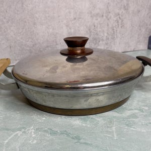 May include: A vintage stainless steel cooking pot with a lid. The pot has a brown wooden handle and a matching knob on the lid. The pot's base has a gold-colored band. The handles are made of metal and wood.