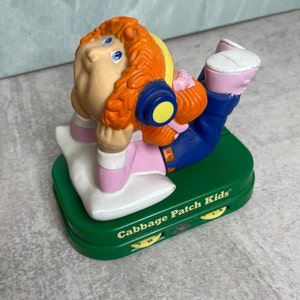 May include: A Cabbage Patch Kids figurine, featuring a doll with orange hair, blue eyes, and headphones, lying on a white pillow. The doll wears a pink shirt and blue pants, resting on a green base with the text "Cabbage Patch Kids".