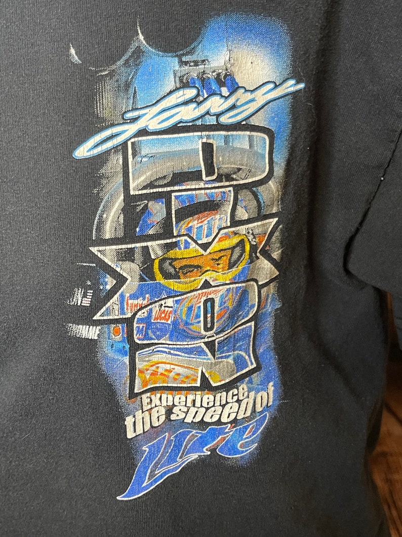 Może przedstawiać: Czarny t-shirt z nadrukiem graficznym zawierającym napis "Lary" u g&oacute;ry i "Experience the speed of Lite" u dołu. Wz&oacute;r zawiera duże białe logo "IROC" nałożone na niebieskawy obraz kierowcy wyścigowego w kasku i goglach ochronnych.