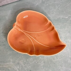 May include: A peach-colored, leaf-shaped ceramic dish. The dish has a glossy finish and features a raised vein design. The dish is designed for serving or display.