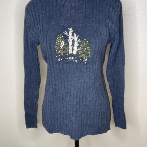 May include: A dark blue ribbed mock neck sweater featuring a central embroidered design of two green pine trees and two white birch trees with small white dots representing falling snow scattered across the chest area of the garment.