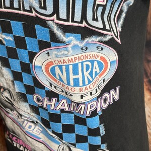 May include: Black t-shirt featuring a graphic of a race car and the text "NHRA Top Fuel Champion 1999." The design includes a checkered flag pattern in blue and black, with the "Exide Racing Team" logo.