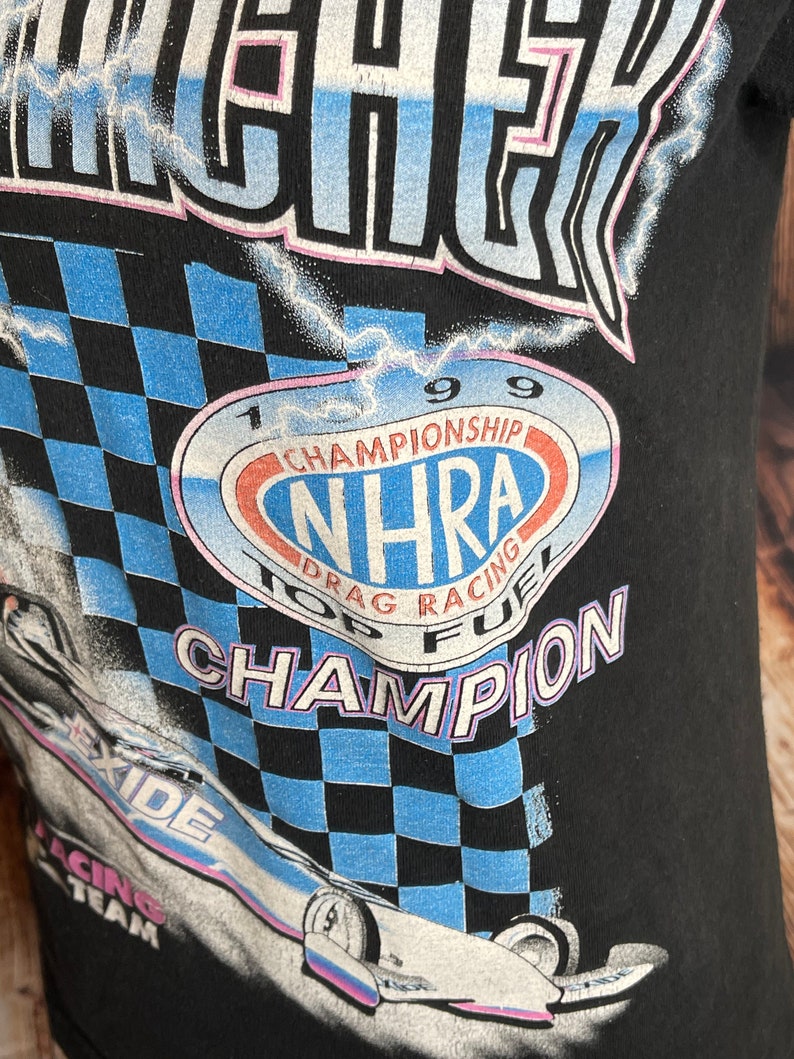 May include: Black t-shirt featuring a graphic of a race car and the text "NHRA Top Fuel Champion 1999." The design includes a checkered flag pattern in blue and black, with the "Exide Racing Team" logo.