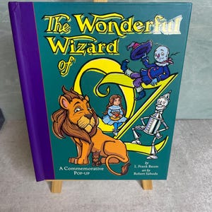 May include: A hardcover pop-up book titled The Wonderful Wizard of Oz by L. Frank Baum with art by Robert Sabuda. The teal cover features illustrations of a lion, a girl, a scarecrow, and a tin man, with a large yellow letter O and Z.
