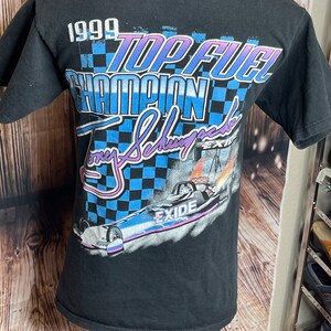 May include: Black t-shirt featuring a graphic design with the text "1999 TOP FUEL CHAMPION Tony Schumacher EXIDE". The design includes a checkered flag pattern and a drag racing car with flames.