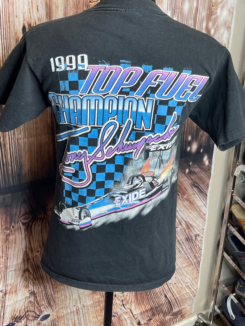 May include: Black t-shirt featuring a graphic design with the text "1999 TOP FUEL CHAMPION Tony Schumacher EXIDE". The design includes a checkered flag pattern and a drag racing car with flames.