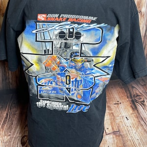 Może przedstawiać: Czarny t-shirt z kr&oacute;tkim rękawem z grafiką na plecach przedstawiającą dragster Don Prudhomme Snake Racing. Projekt zawiera tekst "Don Prudhomme Snake Racing", "Larry", "Miller Lite", "Lucas Oil" oraz "Experience the Speed of Lite".