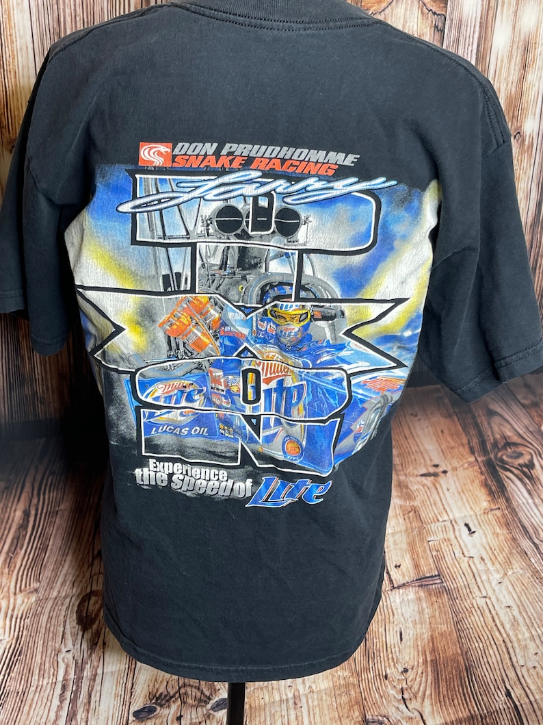 Może przedstawiać: Czarny t-shirt z kr&oacute;tkim rękawem z grafiką na plecach przedstawiającą dragster Don Prudhomme Snake Racing. Projekt zawiera tekst "Don Prudhomme Snake Racing", "Larry", "Miller Lite", "Lucas Oil" oraz "Experience the Speed of Lite".