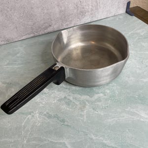 May include: A small, aluminum saucepan with a black handle. The pan has a rounded shape and a pouring spout. The handle is attached with a single rivet. The pan is sitting on a light green and white marble-patterned surface.