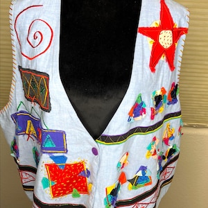 May include: Light blue vest with colorful, abstract patches and a large red star applique. The vest features a V-neck, button closure, and embroidered details in red, orange, and black. The vest is displayed on a black mannequin.