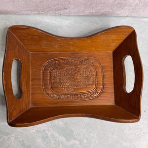 May include: A rectangular wooden tray with a carved central design and two side handles. The tray is a medium brown color and features the text "Give Us This Day Our Daily Bread" in an oval design.