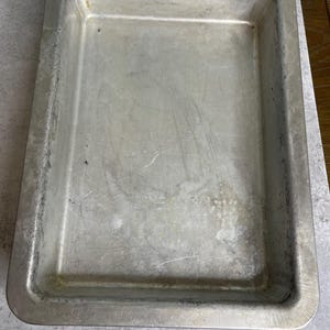 May include: A rectangular, well-used, aluminum baking pan. The pan has a slightly raised edge and shows signs of wear and tear, with scratches and discoloration across the surface. The pan is empty and ready for use.