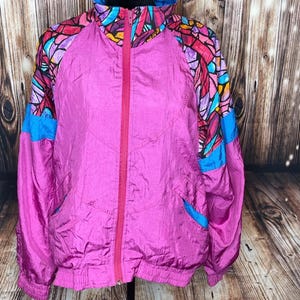 May include: A vibrant pink windbreaker jacket featuring a high collar and a full-length front zipper. The shoulders and upper sleeves display a colorful stained-glass pattern in shades of purple, blue, yellow, and black, with blue accents.