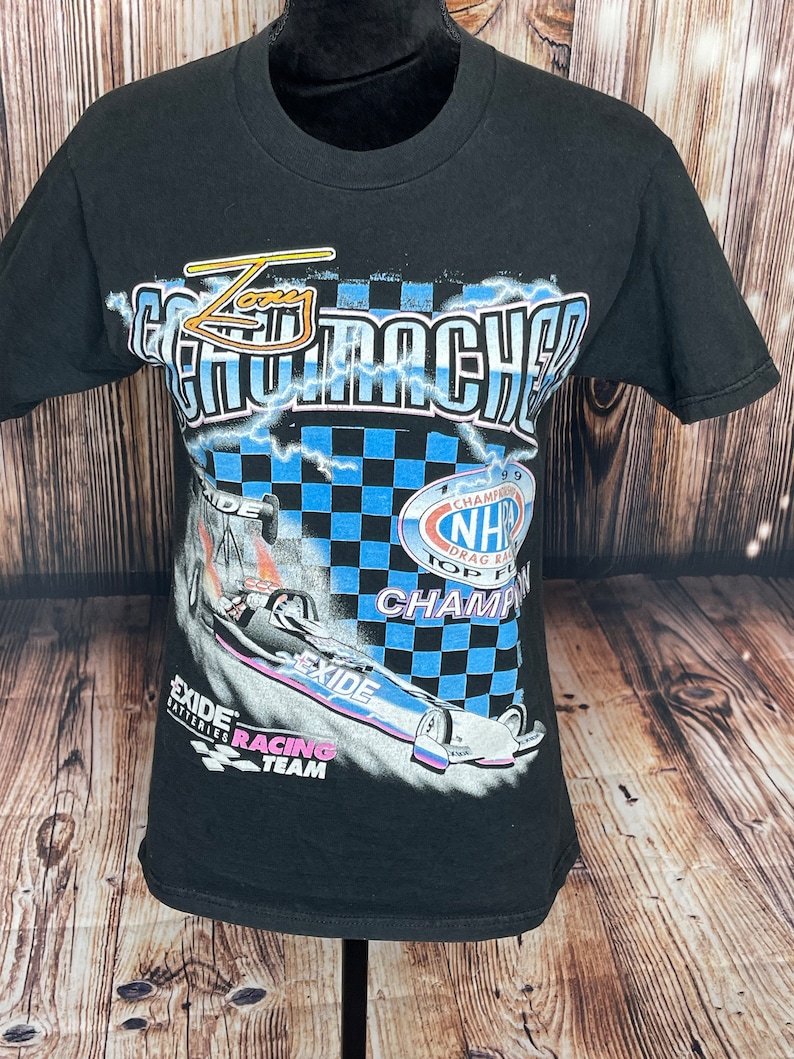 May include: Black t-shirt featuring a drag racing design. The graphic includes the words "Zany Go-Machers" and "Exide Batteries Racing Team". The design also features a race car, lightning bolts, and a checkered flag. The shirt has short sleeves and a crew neck.