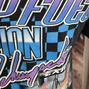 May include: Black t-shirt with a racing-themed graphic. The design features the words "Speed Fuel" in a gradient blue and purple font, along with a checkered pattern and other racing-related imagery. The shirt has a distressed, vintage look.