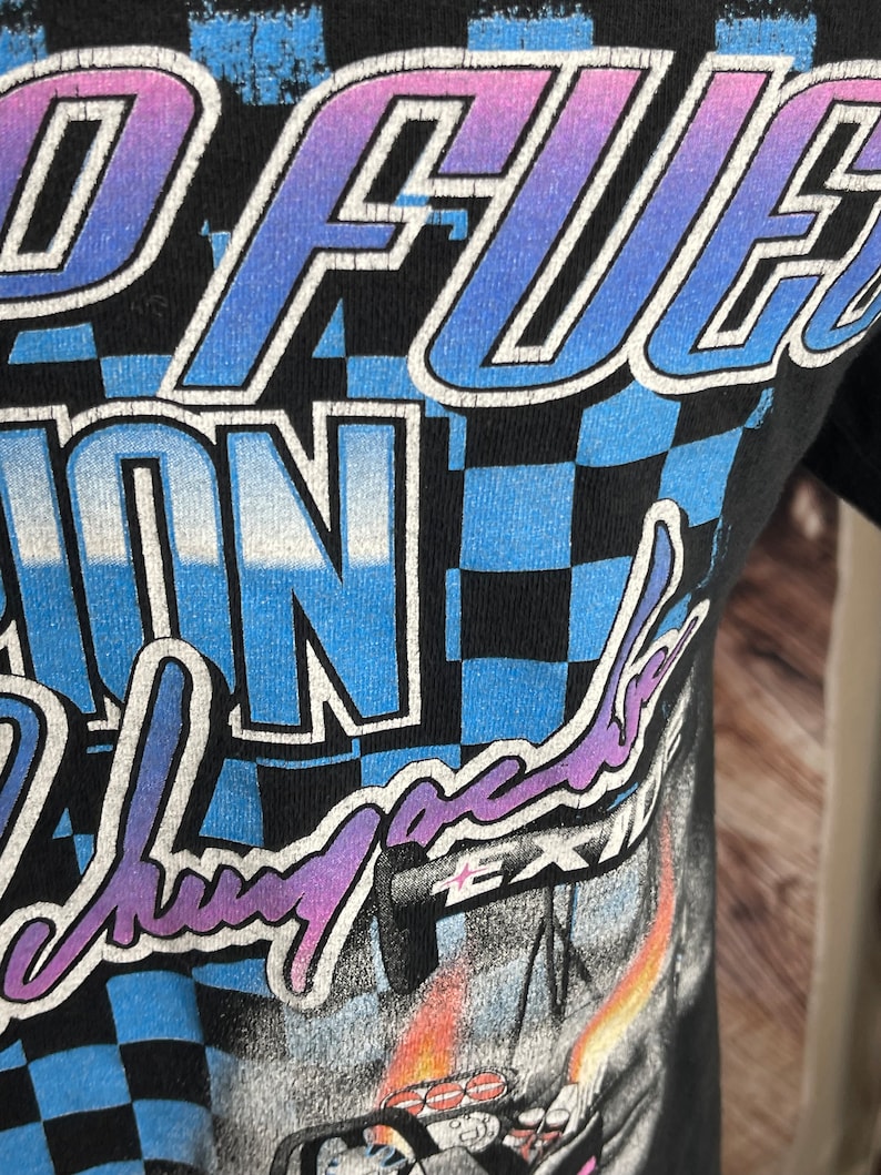 May include: Black t-shirt with a racing-themed graphic. The design features the words "Speed Fuel" in a gradient blue and purple font, along with a checkered pattern and other racing-related imagery. The shirt has a distressed, vintage look.