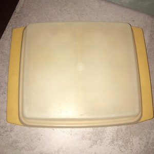 May include: A vintage, rectangular Tupperware container with a pale yellow lid and base. The lid is slightly recessed and fits snugly into the base, which has extended edges. The container is made of durable plastic.