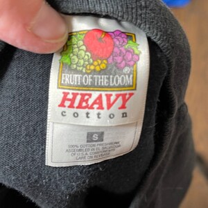 May include: Close-up of a black t-shirt tag. The tag features the "Fruit of the Loom" logo with a red apple, grapes, and other fruit. The tag reads "HEAVY cotton" and "S". The shirt is made of 100% cotton.